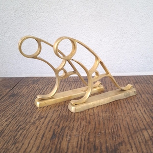 Może przedstawiać: Dwa mosiężne, dekoracyjne wsporniki półek. Każdy wspornik ma prostokątną podstawę, łukowate podparcie i okrągłą górę. Wsporniki są prezentowane na drewnianej powierzchni.