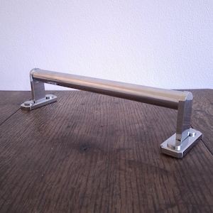 May include: A stainless steel towel bar with a rectangular design. The bar is mounted on two rectangular bases. The bar is displayed on a dark brown wooden surface.