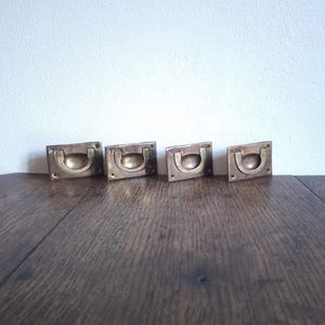 May include: Four antique brass drawer pulls with a rectangular backplate and a curved handle. The pulls are arranged in a row on a dark wooden surface, with a white wall in the background. These vintage hardware pieces are ideal for furniture restoration.