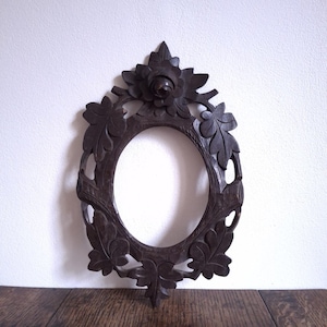 Op de afbeelding: Donkerbruine, ovaalvormige houten lijst met ingewikkelde bloemen- en bladsnijwerk. De lijst heeft een centrale roos en omringend gebladerte, waardoor een decoratieve rand ontstaat. De lijst is ongeveer 30 cm hoog en 23 cm breed, geschikt voor een portret of kunstwerk.