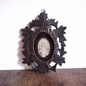 Puede incluir: Un retrato enmarcado ovalado de color marrón oscuro con un diseño de hojas talladas. El retrato presenta una imagen en escala de grises de una persona con un halo y flores. El marco es de madera y tiene un gancho para colgar.