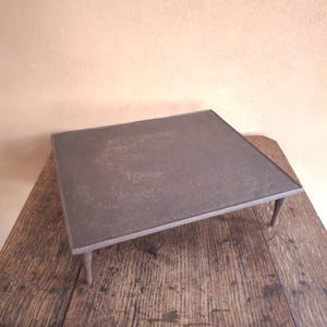 May include: A square, dark grey metal stand with a flat, textured top surface and four slender, tapered legs. The metal has a weathered, matte finish with visible variations in tone across the surface, providing a sturdy and elevated platform.
