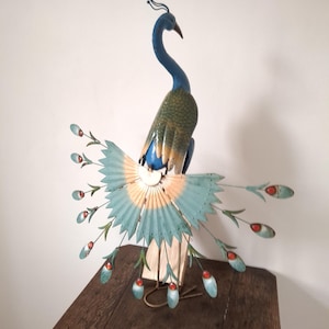 Op de afbeelding: Een decoratieve metalen pauwsculptuur met een blauw lichaam, groene en blauwe vleugels en een waaierachtige staart. De staart heeft blauwe en rode accenten. De sculptuur wordt tentoongesteld op een houten oppervlak.