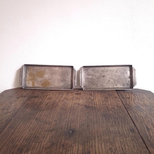 Könnte beinhalten: Zwei rechteckige, silberfarbene Tabletts mit kleinen Griffen an jedem Ende. Die Tabletts stehen nebeneinander auf einer dunklen Holzoberfläche. Die Tabletts weisen Gebrauchsspuren und Alter auf.