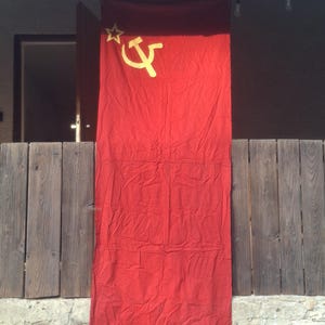Pode incluir: Uma bandeira retangular de tecido vermelho com um símbolo de martelo e foice de cor dourada posicionado no canto superior esquerdo, acompanhado por uma estrela dourada de cinco pontas localizada acima do emblema.
