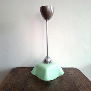 May include: An Art Deco-style ceiling light fixture. It features a metallic, cone-shaped top, a slender, silver-colored stem, and a square, light green glass shade. The fixture is set on a dark wooden table against a white wall.