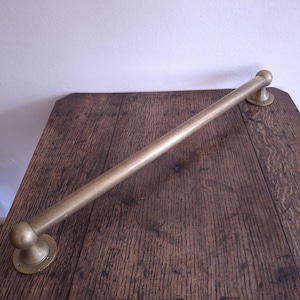 May include: A brass-colored towel bar with a cylindrical design. The bar is mounted on two circular plates with a rounded end on each side. The bar is approximately 24 inches long.