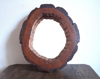 Puede incluir: Un espejo redondo con un marco de madera natural. El marco está hecho de una sección transversal de un tronco de árbol, mostrando la veta y la textura de la madera. El espejo está contra una pared blanca y sobre una mesa de madera.