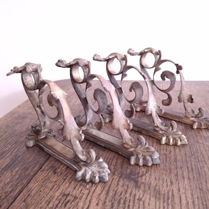 May include: Four antique metal fireplace andirons with ornate, scrolling designs. The andirons have a silver-toned finish and are arranged on a wooden surface. Each andiron features a decorative base and a curved handle.