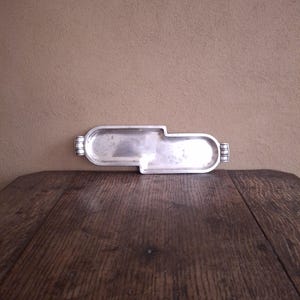Könnte beinhalten: Ein silberfarbenes Serviertablett aus Metall mit einer länglichen, gestuften rechteckigen Form und abgerundeten Enden. Das Tablett verfügt über zwei dekorative Griffe an jeder Seite, die aus drei horizontalen Bändern bestehen, und eine glatte Oberfläche.