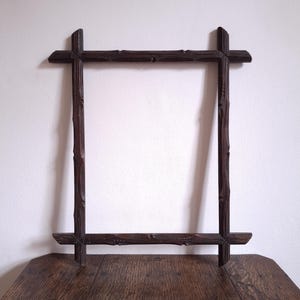 Può includere: Una cornice in legno marrone scuro di forma rettangolare. La cornice ha un design rustico, con angoli e lati a forma di croce. La cornice è appoggiata su un tavolo di legno contro un muro bianco. Dimensioni: circa 40 cm x 50 cm.