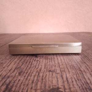 Könnte beinhalten: Ein rechteckiges Zigarettenetui aus goldfarbenem Metall mit einer glatten Oberfläche und einem strukturierten Kreuzschraffurmuster an den Seitenkanten. Das Etui verfügt über einen dünnen, horizontalen Metallverschluss an der Vorderkante.