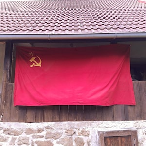 Può includere: Una bandiera rettangolare rossa con un simbolo di martello e falce color oro posizionato nell'angolo in alto a sinistra, accompagnato da una piccola stella dorata a cinque punte situata sopra l'emblema incrociato.