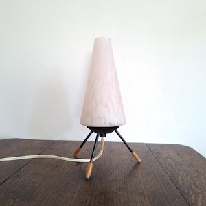 Peut inclure: Une lampe de table vintage avec un abat-jour conique en verre rose et blanc moucheté. La lampe a une base en métal noir avec trois pieds et des embouts en bois. Un cordon électrique blanc s'étend de la base.