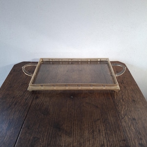 Könnte beinhalten: Ein rechteckiges Serviertablett aus Glas mit einem goldenen Metallrahmen und zwei Griffen. Das Tablett steht auf einer Holzoberfläche.