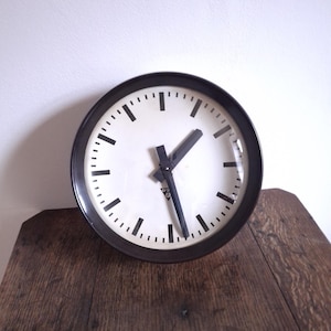 Può includere: Orologio da parete rotondo con cornice nera e quadrante bianco. L'orologio presenta lancette e indicatori delle ore neri. L'orologio è su una superficie di legno.