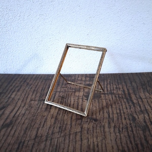 Op de afbeelding: Een gouden metalen fotolijst met een decoratieve rand. De lijst staat rechtop op een houten oppervlak.