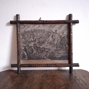 May include: A framed monochrome print of a historical battle scene. The print is set in a dark wooden frame with a cross design at each corner. The text at the bottom of the print is unclear.