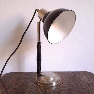 May include: An antique desk lamp with a black shade and a silver-colored base. The lamp has a black cord and a wooden handle. The lamp is on a wooden surface.