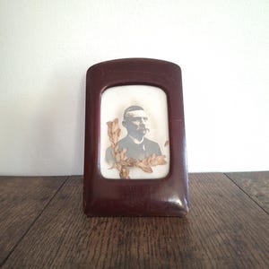 Puede incluir: Un marco de fotos de madera marrón oscuro con un retrato en blanco y negro de un hombre con traje. El marco, con la parte superior curvada, descansa sobre una superficie de madera. Hojas secas adornan el frente del retrato.