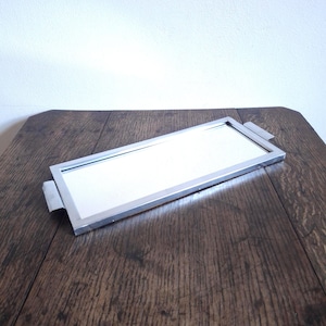 May include: A rectangular silver-toned mirror tray with a mirrored surface and small handles on each end. The tray is displayed on a dark wooden surface, reflecting the light. The tray is a decorative item.