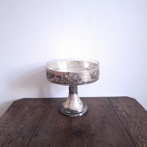 May include: A silver-plated pedestal serving dish with a clear glass top. The base features an ornate design. The dish is placed on a dark wooden table against a white wall. A vintage serving piece.