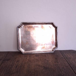 May include: A rectangular silver-toned metal tray with rounded corners and a raised, scalloped edge. The surface of the tray shows signs of wear and tarnishing, with visible patches of copper-coloured oxidation across the centre and edges.
