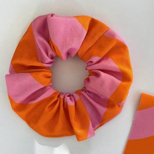 May include: A fabric hair scrunchie featuring a repeating wavy pattern of bright orange and light pink stripes. The scrunchie has a gathered, pleated texture around the elastic centre, and a matching piece of fabric is visible in the corner.