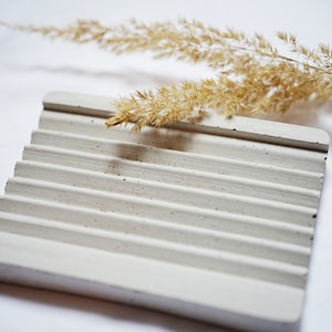 Può includere: Un portasapone in cemento bianco con una superficie testurizzata e un design a coste. Il portasapone è appoggiato su una superficie bianca con uno stelo d'erba secca sopra.