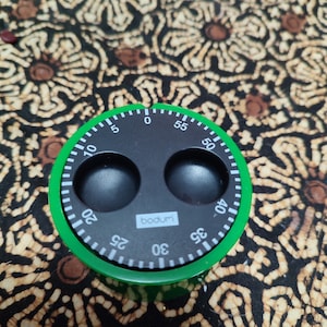 Puede incluir: Un temporizador de cocina verde con una cara negra y dos botones negros. El temporizador tiene una esfera blanca con números negros del 0 al 60. El temporizador está etiquetado como "bodum".