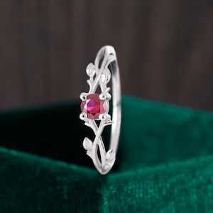 May include: A silver ring with a red gemstone set in a delicate floral design. The ring is on a green velvet background.