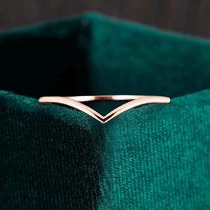 May include: A rose gold V-shaped ring, set against a dark green velvet background. The ring's design is a simple, elegant chevron shape. The ring is a delicate piece of jewelry.