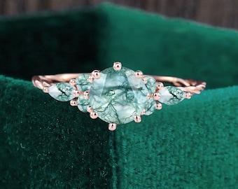May include: A rose gold ring featuring a large, round, moss agate gemstone with green inclusions. The ring has two smaller pear-shaped moss agate stones on either side of the main stone, and a twisted band.