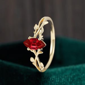 Può includere: Un anello d'oro con una vivace pietra rossa a taglio marquise. La fascia e l'incastonatura dell'anello sono decorate con delicati dettagli a forma di foglie, creando un design elegante e ispirato alla natura. Sfondo verde scuro.