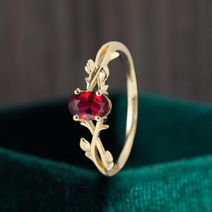 May include: A gold ring with an oval, deep red gemstone. The ring's band has a delicate, floral design with leaves and stems. The gemstone is the focal point, set in a gold setting.