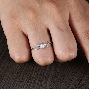 Op de afbeelding: Een zilveren ring met een delicaat takontwerp en een rond maansteencentrum. De ring wordt gedragen aan een vinger.