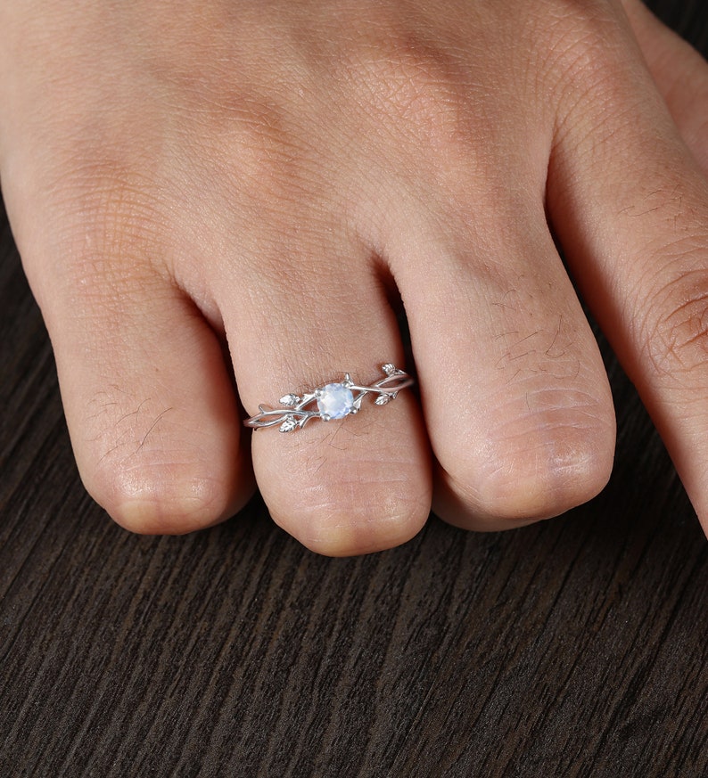 Op de afbeelding: Een zilveren ring met een delicaat takontwerp en een rond maansteencentrum. De ring wordt gedragen aan een vinger.