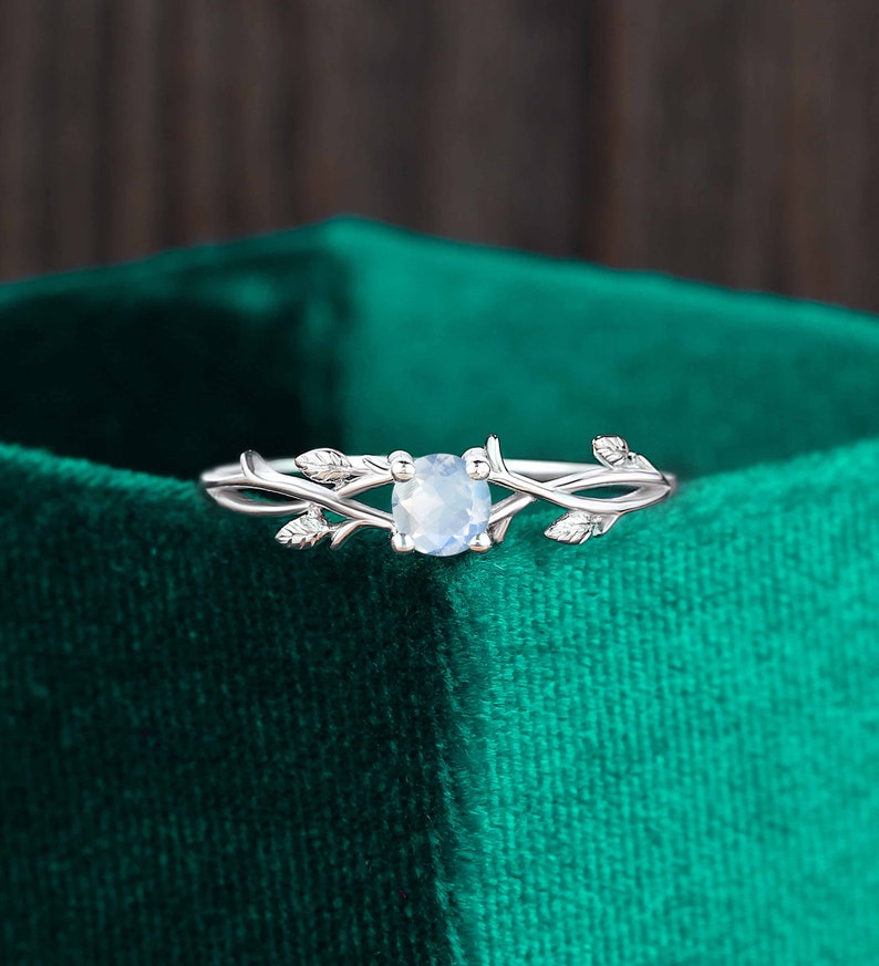 Op de afbeelding: Een zilveren ring met een delicaat takontwerp en een heldere ovale edelsteen. De ring ligt op een groene fluwelen achtergrond.