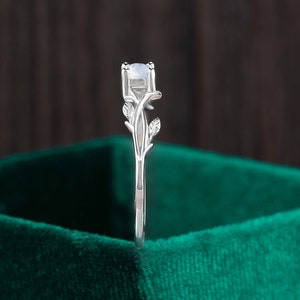 Op de afbeelding: Een zilveren ring met een delicaat takontwerp en een ronde, witte edelsteen. De ring is geplaatst tegen een groene fluwelen achtergrond.