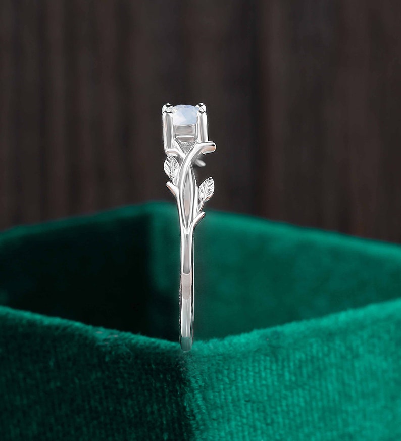 Op de afbeelding: Een zilveren ring met een delicaat takontwerp en een ronde, witte edelsteen. De ring is geplaatst tegen een groene fluwelen achtergrond.
