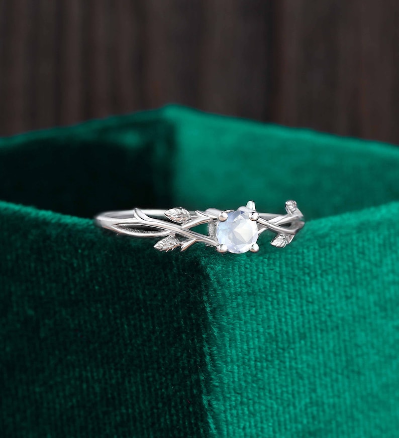 Op de afbeelding: Een zilveren ring met een delicaat takontwerp en een ronde, heldere edelsteen. De ring ligt op een groene fluwelen achtergrond.