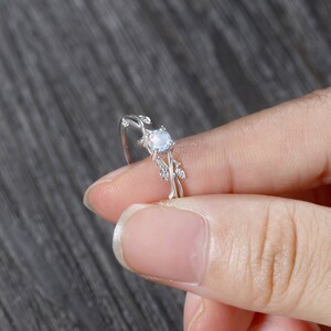 Op de afbeelding: Een zilveren ring met een delicaat, takvormig ontwerp met een ronde, heldere edelsteen. De ring zit op een vinger.