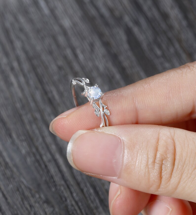 Op de afbeelding: Een zilveren ring met een delicaat, takvormig ontwerp met een ronde, heldere edelsteen. De ring zit op een vinger.