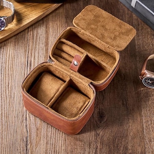 May include: A brown leather watch box with a soft brown interior. The box has two compartments for watches and a smaller compartment for jewelry.