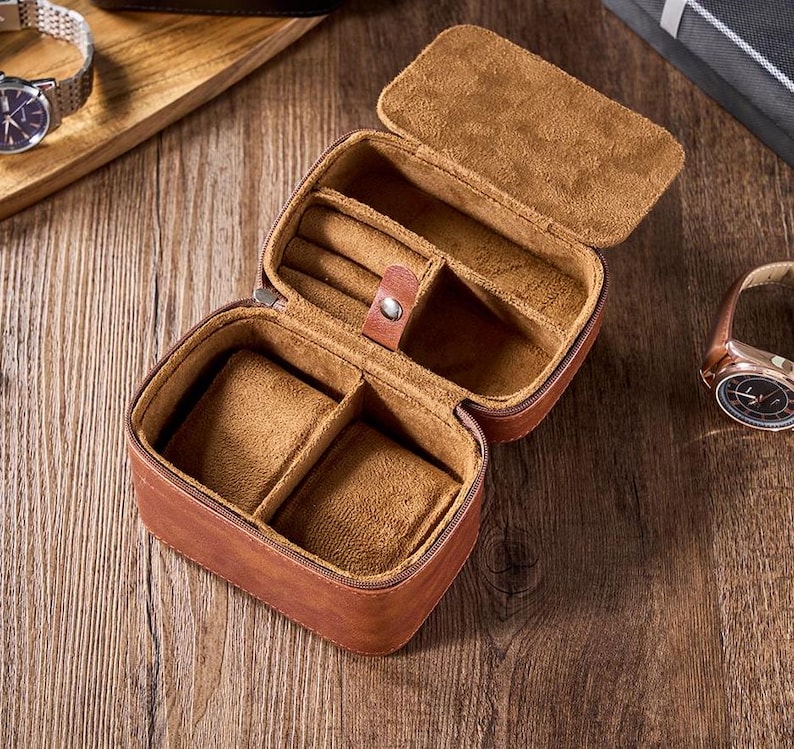 May include: A brown leather watch box with a soft brown interior. The box has two compartments for watches and a smaller compartment for jewelry.
