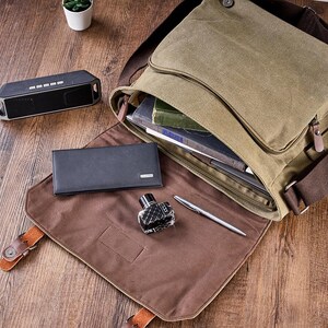 May include: An olive green canvas messenger bag with a brown interior flap, featuring a black rectangular wallet, a small glass ink bottle, and a silver pen placed on the open flap, with a black portable speaker nearby.
