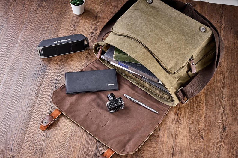 May include: An olive green canvas messenger bag with a brown interior flap, featuring a black rectangular wallet, a small glass ink bottle, and a silver pen placed on the open flap, with a black portable speaker nearby.