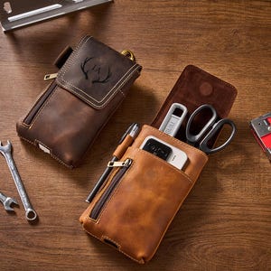 Puede incluir: Dos bolsas de cuero marrón, una con un diseño de cornamenta de ciervo, y una bolsa de cuero bronceado con cremallera. También se ven una grapadora roja, llaves, tijeras y un teléfono.