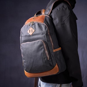 Puede incluir: Una mochila de lona negra con detalles de cuero marrón. La mochila tiene un compartimento principal grande, un bolsillo frontal más pequeño y correas de hombro ajustables.