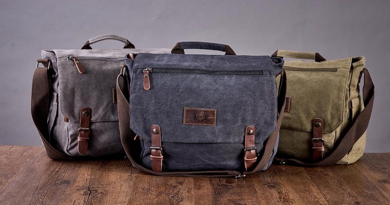 May include: Three canvas messenger bags in gray, dark blue, and olive green. Each bag features a top handle, a wide shoulder strap, a zippered front pocket, and a flap closure secured by two brown leather straps with metal buckles and snap buttons.